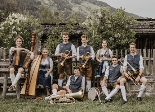 The musicians of Zillertaler Tanzlmusig pose with their instruments in front of a rustic wooden chalet.